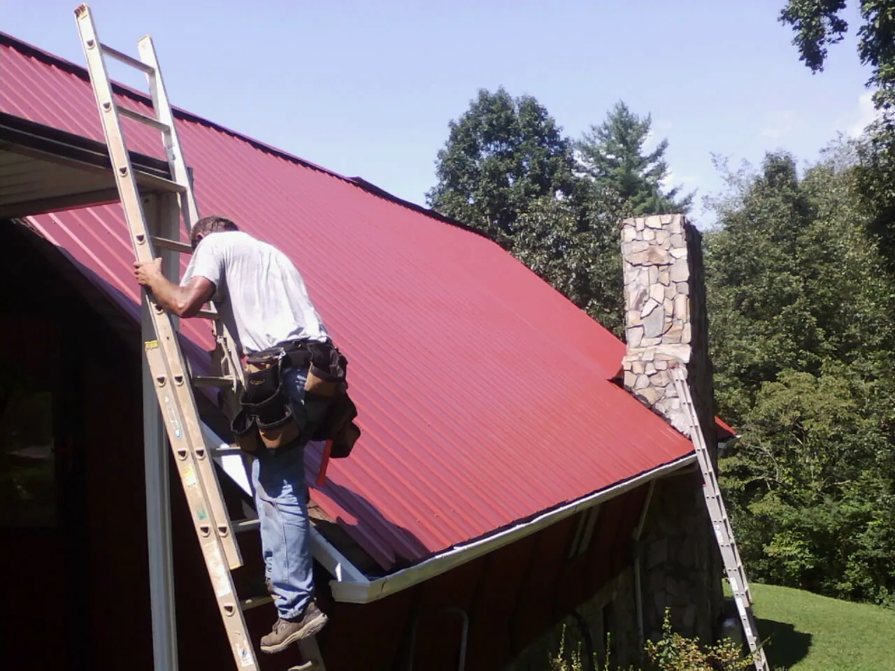 Roofer on a ladder beside a red metal roof and stone chimney — Soffit & Fascia Repair by Roofing Brothers in Lissie
